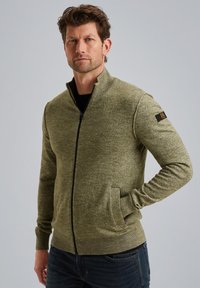 Grøn lynlåstrøje med høj krave, strikket tekstur, ribstrikkede manchetter og kant, samt sidelommer. Har et logo på venstre ærme.