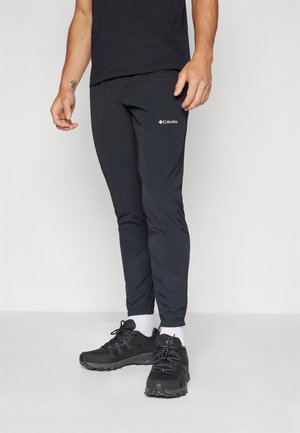 Homme portant un pantalon de jogging noir de la marque Columbia, des chaussures de sport noires, des chaussettes blanches et un t-shirt noir, debout sur un fond uni.