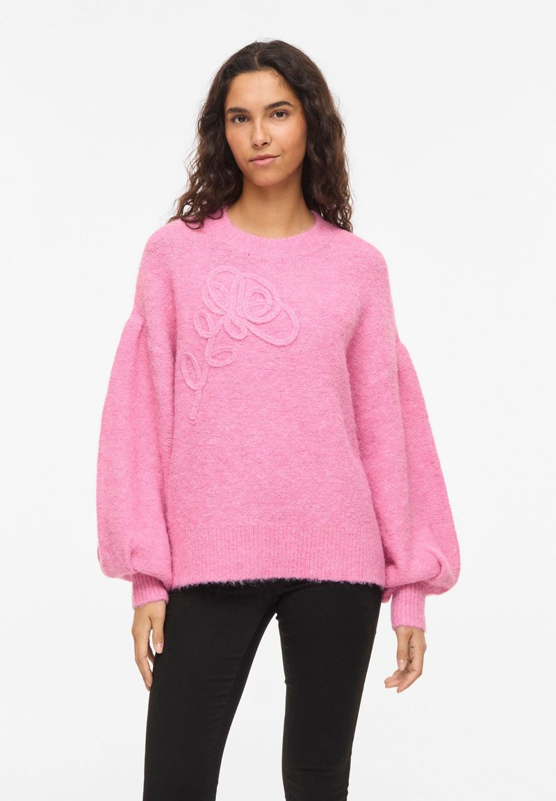 Pull rose en tricot avec manches bouffantes et détail de broderie florale texturée sur le devant. Taille et poignets côtelés.