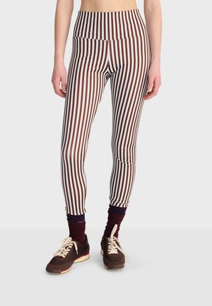 Personne portant des leggings marron et blanc à rayures verticales taille haute, des chaussettes foncées et des baskets marron avec des lacets crème sur un fond uni.