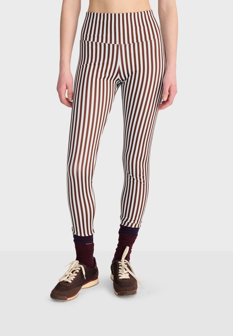 Personne portant des leggings marron et blanc à rayures verticales taille haute, des chaussettes foncées et des baskets marron avec des lacets crème sur un fond uni.