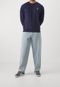 Felpa navy con scollatura rotonda e piccolo logo, abbinata a jeans a gamba larga blu chiaro e sneaker grigie con dettagli bianchi.