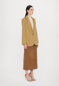 Raidallinen sinapinvärinen bleiseri, beige paita, ruskea mokkaneulospituinen hame ja ihonväriset remmikengät. Bleiserissä on yksi nappi ja leikkaukset.