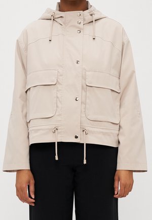 Modèle portant une veste à capuche beige clair avec des boutons-pression, des cordons de serrage et de grandes poches à rabat, par-dessus un pantalon noir.