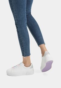 White sneaker with textured sole, pastel laces, and iridescent heel accent. Worn with cropped blue denim, revealing frayed hem.