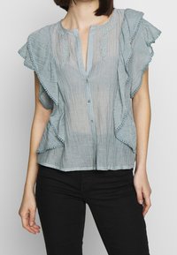 Vaaleansininen, teksturoitu paita, jossa on röyhelöhihoja, kirjontaa kaula-aukon ympärillä ja nappikiinnitys. Yhdistetty mustiin housuihin.