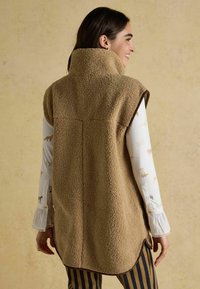 Beige Fleece-Weste mit hohem Kragen, abgerundetem Saum und dunkelbraunem Besatz. Getragen über einem Langarmshirt mit Tiermotiven.