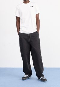 Vit bomullströja med liten svart logotyp, kombinerad med löst sittande svarta denimbyxor och svarta sneakers med vita detaljer.