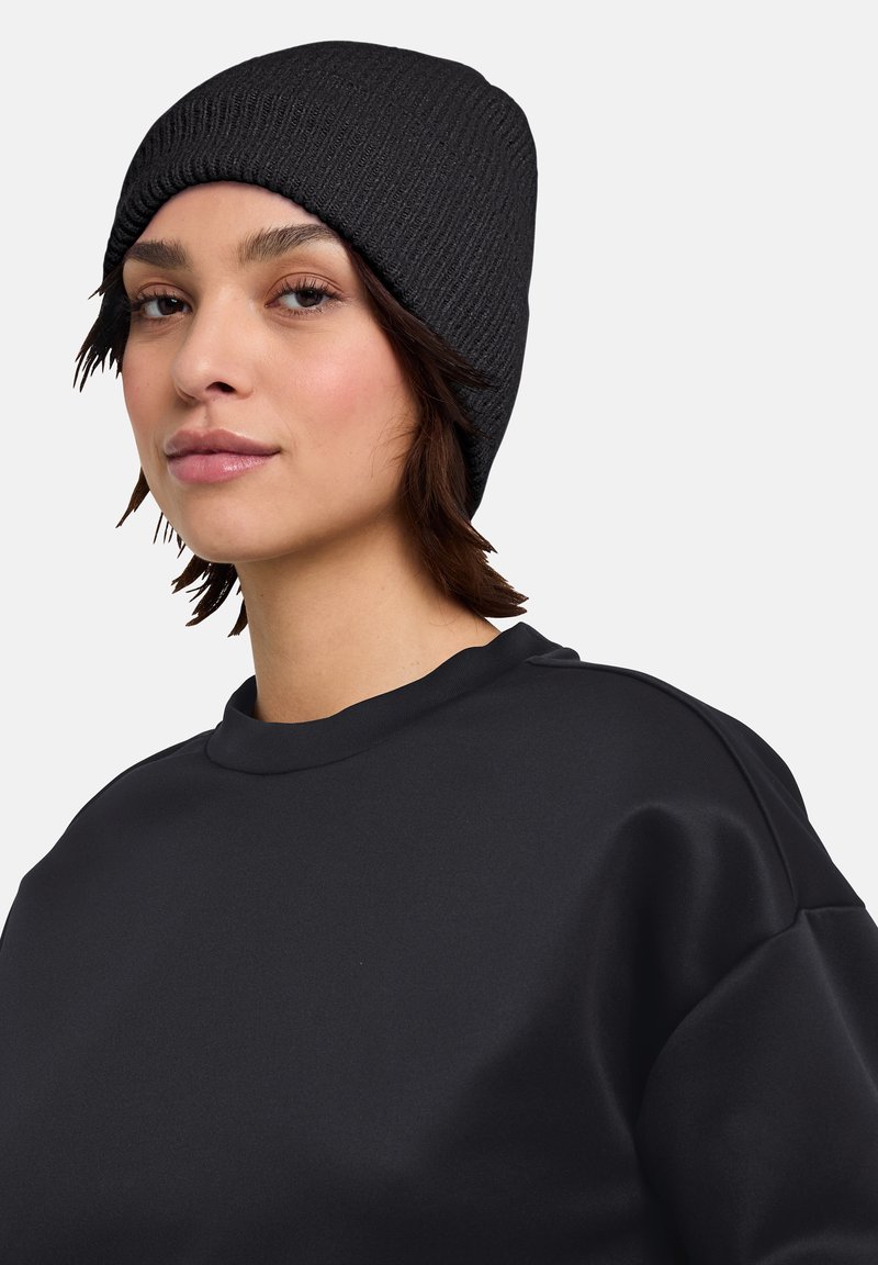 Beanie nera di maglia con tessuto testurizzato, indossata leggermente scivolata, abbinata a una felpa nera a collo rotondo con tessuto liscio e vestibilità rilassata.