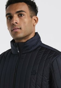 Man met kort krullend haar in een zwarte doorgestikte jas met een staande kraag en een klein geborduurd pinguïnlogo op de borst.