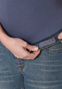 Džíny z lehce modrého denimu s vybledlou texturou. Pas zdobí šedá nastavitelná záložka s knoflíkem, kterou drží ruka.