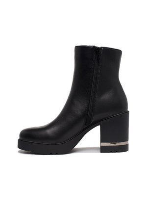 Bottine en cuir noir avec fermeture éclair latérale, talon bloc imposant et accent métallique près de la semelle sur fond blanc.