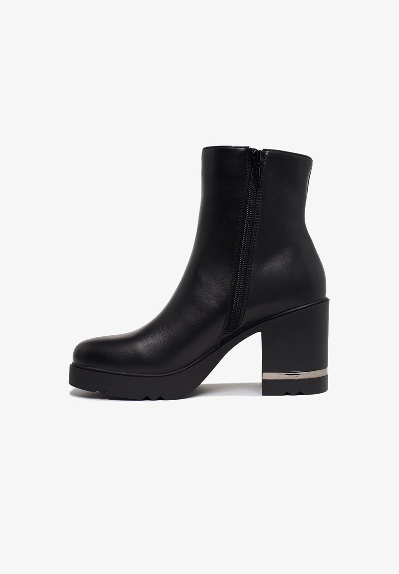 Bottine en cuir noir avec fermeture éclair latérale, talon bloc imposant et accent métallique près de la semelle sur fond blanc.