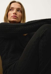 Svart stickad tröja och matchande byxor med flätmönster. Mjuk textur, lös passform, framträdande långa ärmar och avslappnad pose.