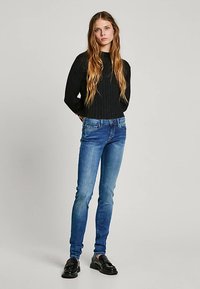 Fekete bordázott pulóver, kék skinny farmer enyhe kopással, és fekete mokaszinok. A modell áll, zsebre tett kézzel, hosszú hajjal.