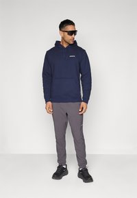 Sweat-shirt à capuche navy avec poche avant, pantalon de jogging gris et chaussures de sport noires. Le sweat-shirt présente un petit logo sur la poitrine.