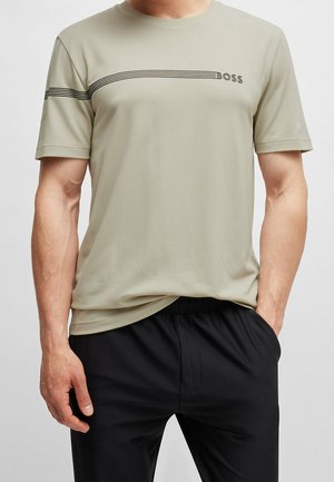 Homme portant un t-shirt à manches courtes beige avec des rayures horizontales noires et le texte "BOSS", associé à un pantalon noir, les mains dans les poches.