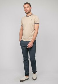 T-shirt beige à manches courtes avec des bordures bleu marine et un petit logo, associé à un pantalon foncé à motifs et des chaussures beiges à enfiler.