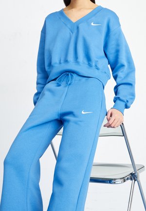Personne portant un pantalon de survêtement Nike bleu et un sweat-shirt à col en V assorti, appuyée sur une chaise moderne en métal et plastique.