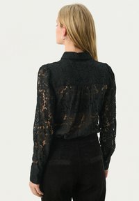 Zwarte kanten blouse met lange doorschijnende mouwen, ingewikkelde bloempatronen en een kraag. De achterkant is gedeeltelijk gevoerd.