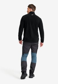 Schwarze Fleecejacke mit hohem Kragen, kombiniert mit schwarz-blauen Farbblock-Hosen. Mit Reißverschlüssen und strukturiertem Stoff.