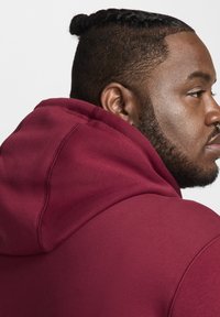 Perfil lateral de un hombre con un moño de trenzas que lleva una sudadera color burdeos contra un fondo blanco.