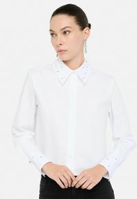 Camicia bianca a maniche lunghe con colletto a punta decorato con borchie trasparenti, apertura frontale con bottoni e polsini impreziositi, abbinata a jeans neri.