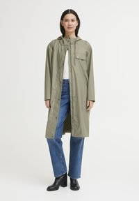 Olivengrøn vandtæt lang frakke med hætte, trykknapper og en forlomme. Kombineret med blå lige ben jeans og sorte hælede støvler.