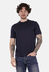 T-shirt a maniche corte blu navy in cotone, con colletto rotondo e un logo discreto sul petto. Sfondo jeans con tatuaggi visibili.