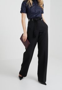 Donna che indossa pantaloni neri a vita alta e larghi con cintura, una blusa corta a maniche navy, tacchi neri, mentre tiene una borsa a tracolla di un viola scuro.