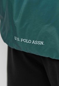 Groene nylon jas met wit "U.S. POLO ASSN." logo aan de onderkant. Beschikt over een gladde textuur, lichtgewicht ontwerp en een ontspannen pasvorm.