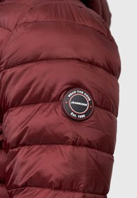 Jachetă puffer bordo cu textură matlasată și o etichetă rotundă cu logo care spune "WEAR THE CODE JEANSCODE Est. 1993." Finisaj din material neted.