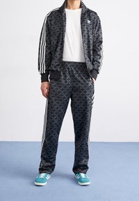 Svart träningsjacka och byxor med ett mönster som har vita tre-stripes detaljer, tillsammans med teal sneakers. Vit T-shirt under.