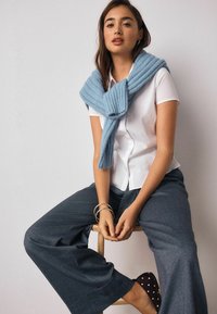 Svetlomodrý rebrovaný pletený sveter prehodený cez bielu košeľu, v kombinácii so sivými širokými nohavicami a topánkami s bodkami. Modelka sedí na stoličke.