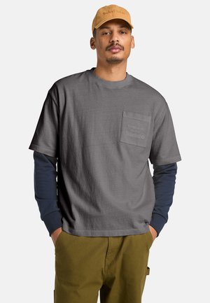 Szara koszulka z krótkim rękawem z przednią kieszenią, ozdobiona subtelnym logo Timberland, nałożona na granatową koszulę z długim rękawem, zestawiona z oliwkowozielonymi spodniami.