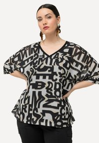 Gennemsigtig sort og beige bluse med overdimensionerede bogstaver i sort. V-hals design, løse ærmer og blød tekstur.