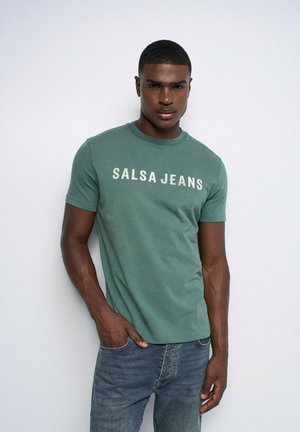 Jeune homme portant un T-shirt vert "SALSA JEANS" et un jean bleu, debout avec une main dans la poche devant un fond blanc.
