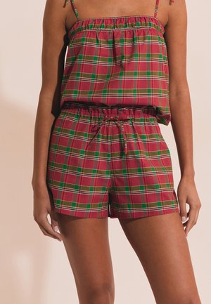 Kobieta ubrana w czerwoną i zieloną koszulkę bez rękawów w kratkę oraz dopasowane szorty ze sznurkiem w pasie na jasnym tle.
