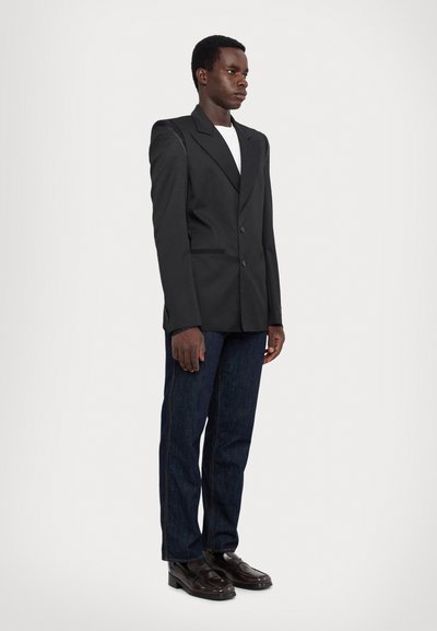 Blazer noir ajusté avec revers crantés, deux boutons et une forme structurée, associé à un jean en denim foncé et des chaussures noires.