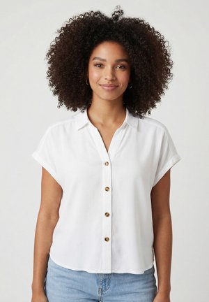 Donna con capelli ricci che indossa una camicia bianca a maniche corte con bottoni e jeans azzurri chiari su uno sfondo semplice.