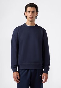 Sudadera navy hecha de tela suave, con cuello redondo, puños acanalados y un pequeño logo en el pecho. Combinada con pantalones a juego.