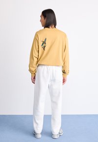 Sweatshirt jaune avec broderie d'oiseau dans le dos, associé à un pantalon de jogging blanc. Le mannequin se tient de dos face à un arrière-plan neutre.