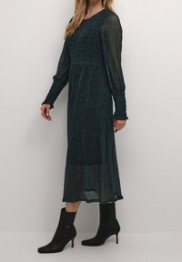 Lång, mörk teal klänning med texturerad, smockad byst och genomskinliga ärmar. Matchad med figursydda svarta ankelskor med spetsig tå.