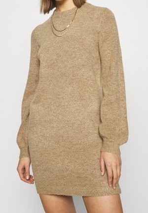 Donna che indossa un vestito a maglia beige a maniche lunghe con maniche a sbuffo e collane in oro a più strati.