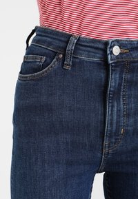 Vaqueros de mezclilla azul oscuro con un diseño ajustado, que cuentan con cinco bolsillos, trabillas para cinturón y un cierre frontal con botón. Textura suave con ligero desvanecimiento.
