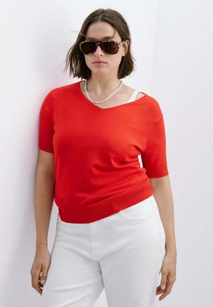 Femme portant des lunettes de soleil écaille, un collier chaîne argenté, un haut rouge à manches courtes et un pantalon blanc, debout contre un mur blanc.