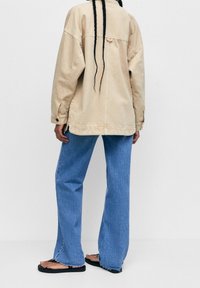 Personne avec de longues tresses portant une veste beige, un jean bleu ample avec des bords effilochés, et des tongs noires, se tenant devant un fond uni.