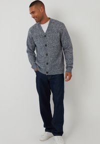 Cardigan grigio a maglia con scollo a V, bottoni frontali e polsini a costine, abbinato a jeans blu scuro e sneaker bianche.