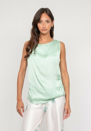 Femme aux longs cheveux bruns portant un haut sans manches en satin vert clair et un pantalon blanc à imprimé floral vert, debout devant un fond clair uni.