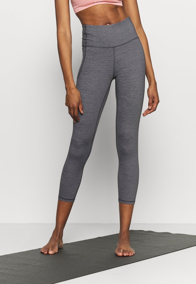 Personne debout pieds nus sur un tapis de yoga, portant un legging gris taille haute et un top court rose sur un fond uni.